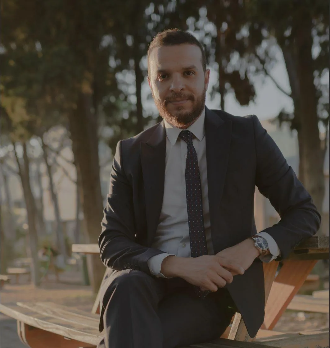 Mohammed Mansour seated outdoors on a wooden bench in a suit