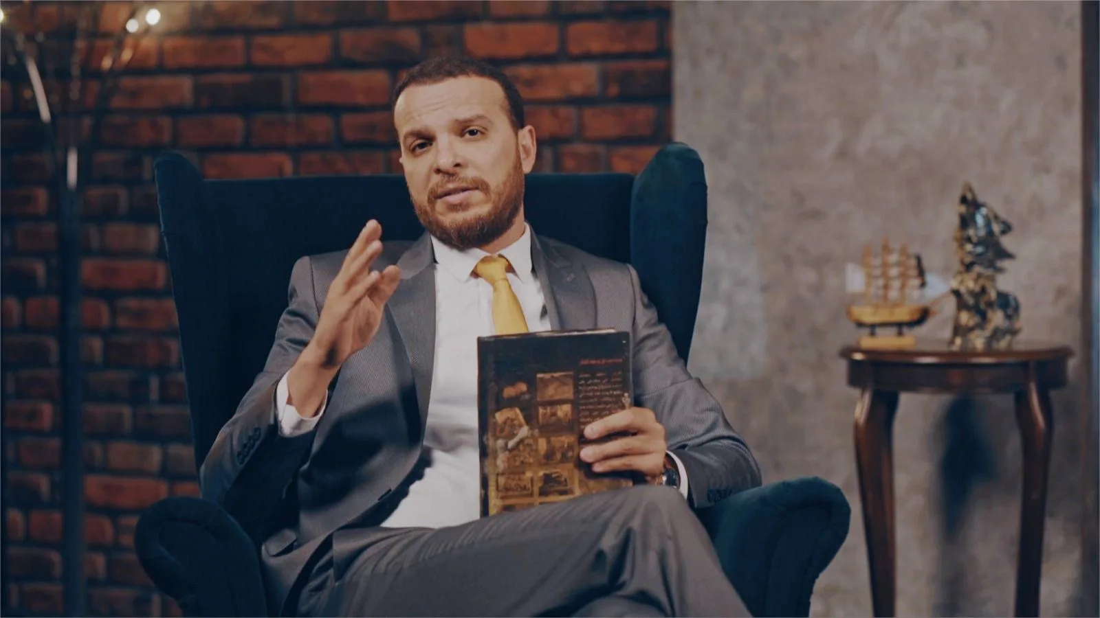 Mohammed Mansour seated in an armchair, holding a book and gesturing as he talks
